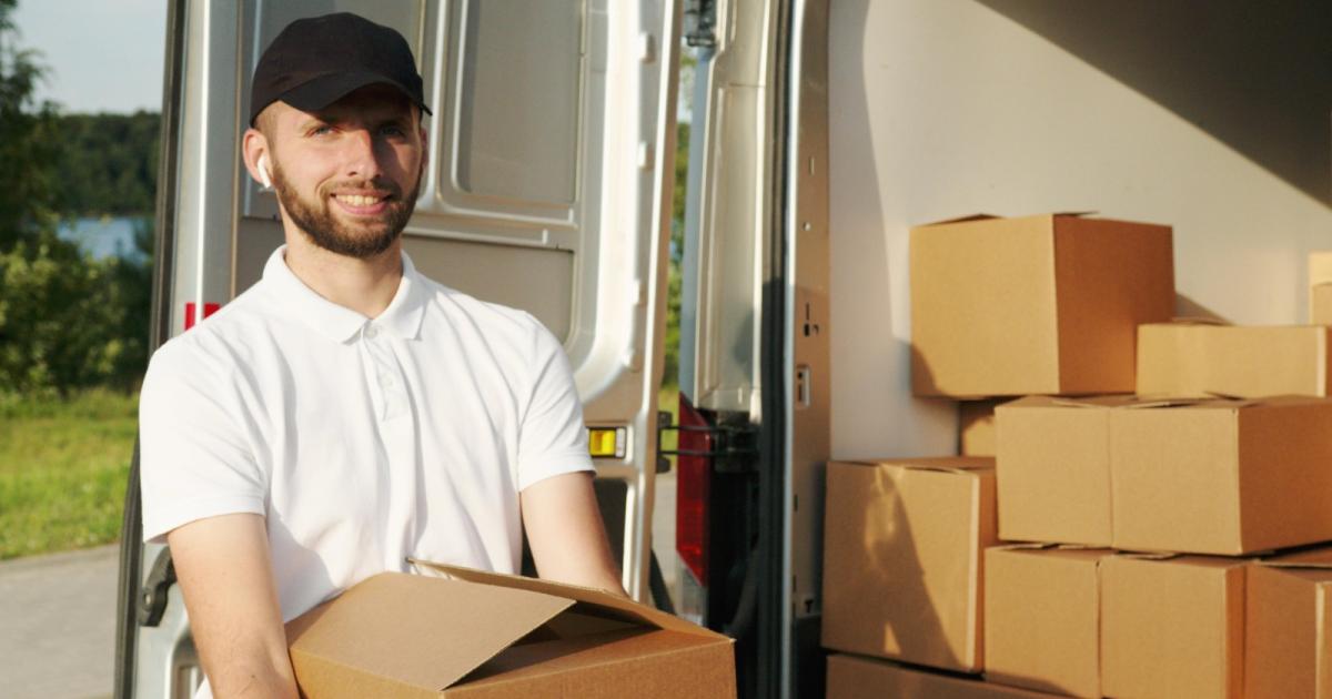 Foto de un un hombre con camiseta blanca y gorra negra alzando una caja sonriendo a la cámara