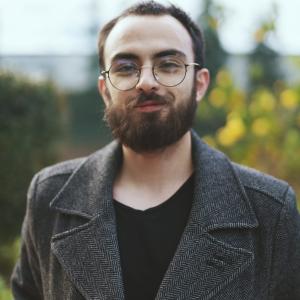 Foto de un hombre con barba y fondo verde con una gabardina gris mirando la cámara