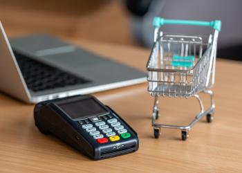 Foto de un datafono con un computador al fondo y al costado un carrito de compras