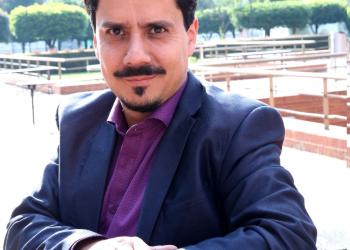 Foto de un hombre blanco con bigote mirando a la cámara, utiliza blazer azul y camiseta vinotinto