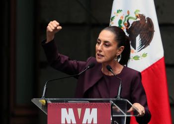 Foto de una mujer con el puño arriba y detrás de ella una bandera de méxico