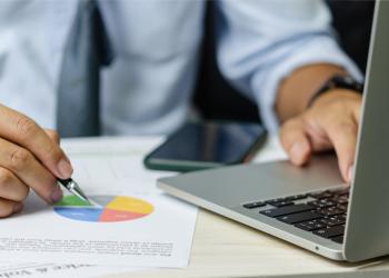 Foto de una persona escribiendo sobre un computador y anotando datos en un papel