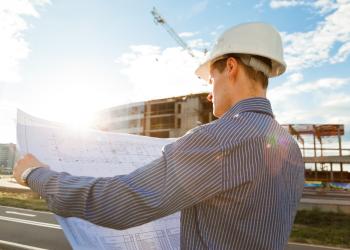 Fotografía de un constructor con camiseta azul sosteniendo en sus manos un plano de construcción y una edificación de fondo