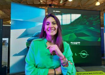 Una mujer con los brazos cruzados mirando a la cámara con una pantalla grande que en el fondo dice Opel