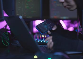representación de hacker, con una computadora al frente y viendo datos en un celular a su costado derecho