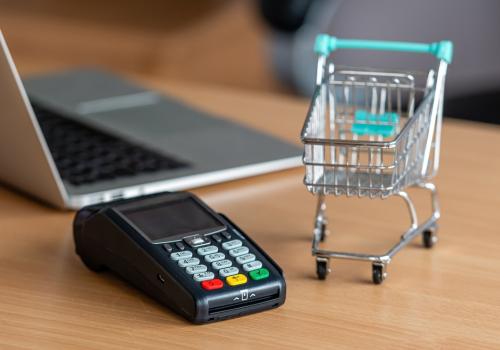 Foto de un datafono con un computador al fondo y al costado un carrito de compras