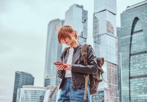 Foto de una mujer con chaqueta roja mirando un celular y unos edificios de fondo