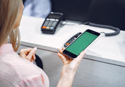 Foto de una mujer portando un celular en la mano con un datafono al fondo