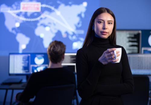 Mujer mirando a la cámara con un fondo de una persona trabajando en un computador y un diseño de continentes arriba de él