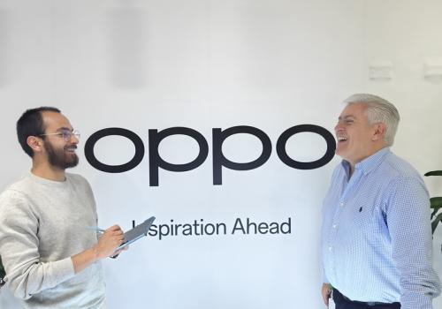 Fotografía de dos personas hablando con un letrero de fondo que dice Oppo