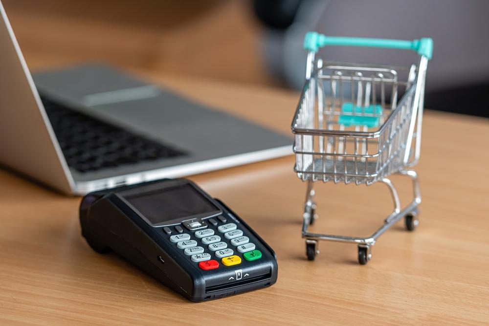 Foto de un datafono con un computador al fondo y al costado un carrito de compras