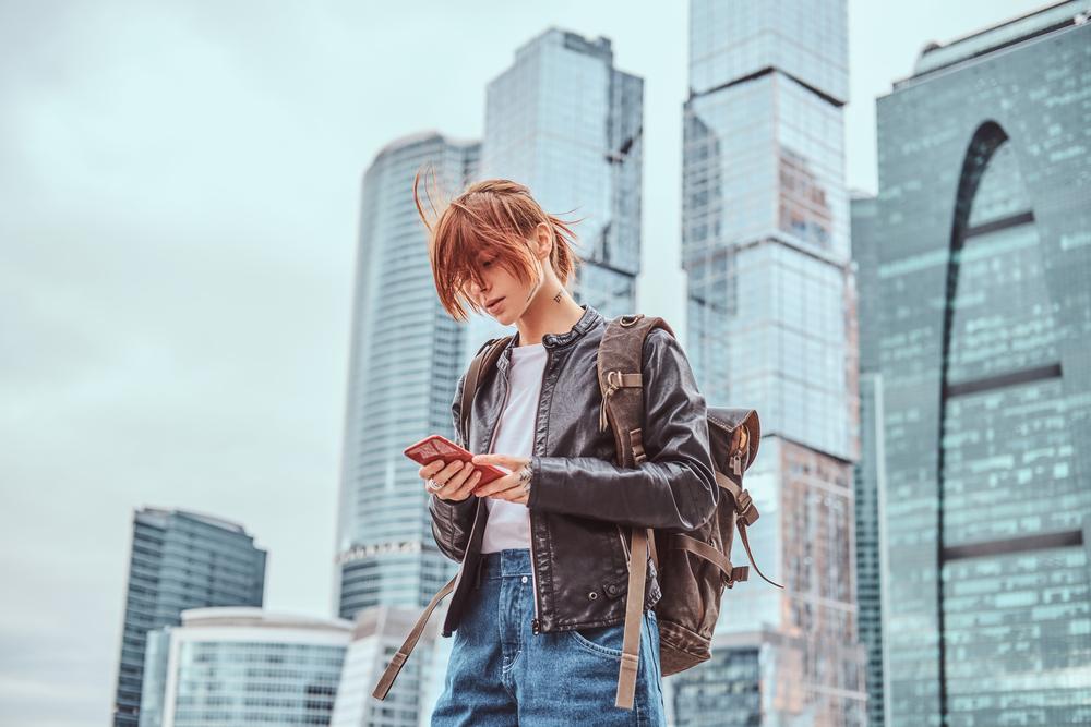 Foto de una mujer con chaqueta roja mirando un celular y unos edificios de fondo