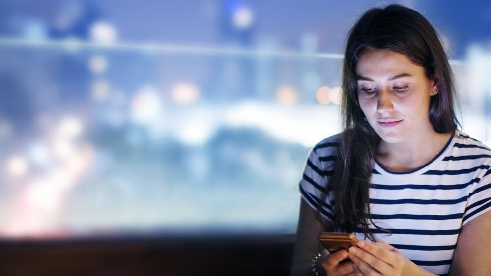 Foto de una mujer mirando el celualr y de fondo una ciudad difuminada