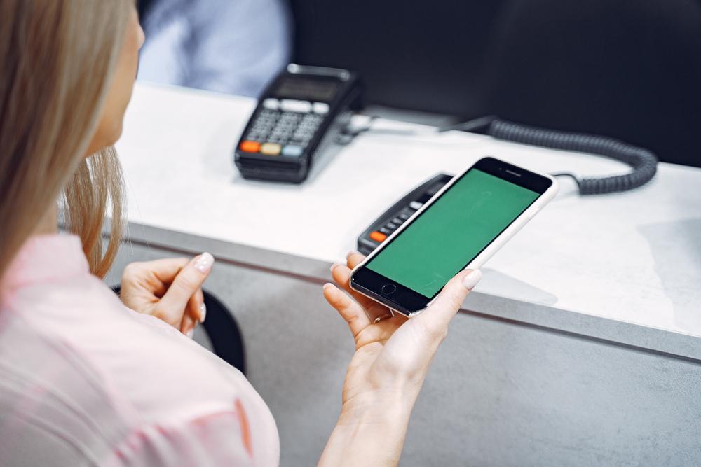 Foto de una mujer portando un celular en la mano con un datafono al fondo