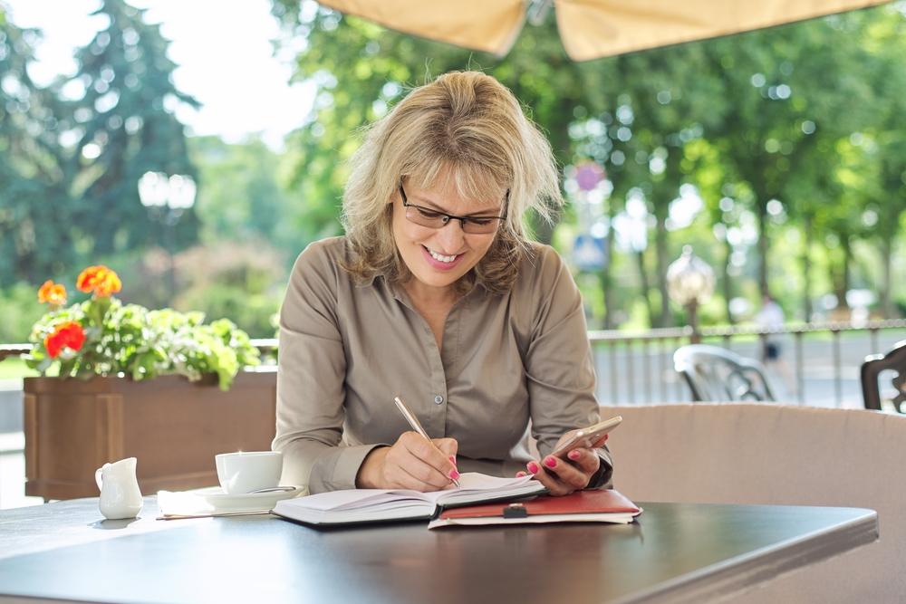 Foto de una mujer rubia anotando algo en un cuaderno con un fondo de arboles