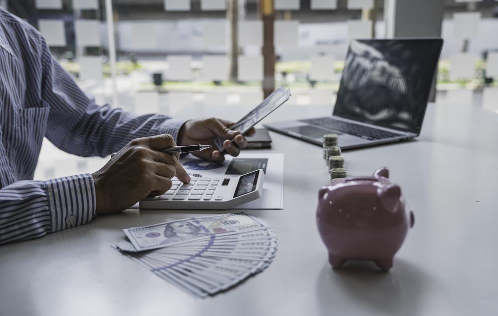 Foto de una persona escribiendo en un computador y al lado un cochinito