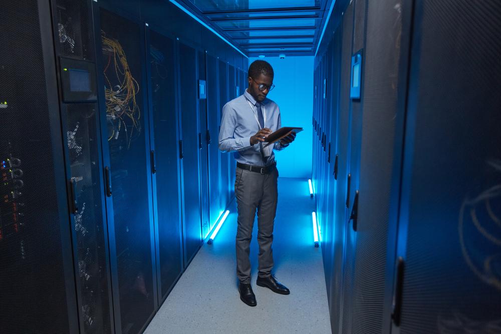 Hombre afroamericano con pantalón gris y camisa azul mirando una tablet en un centro de datos