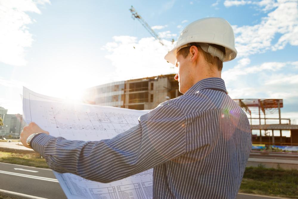 Fotografía de un constructor con camiseta azul sosteniendo en sus manos un plano de construcción y una edificación de fondo