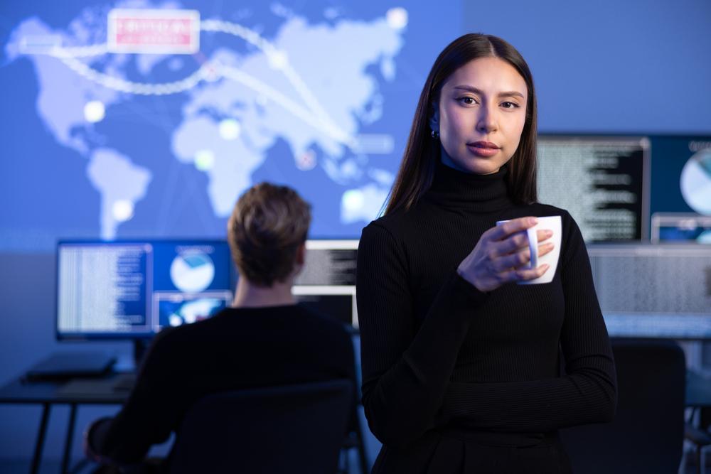 Mujer mirando a la cámara con un fondo de una persona trabajando en un computador y un diseño de continentes arriba de él