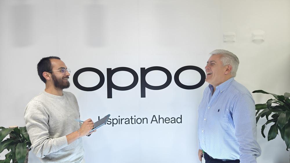 Fotografía de dos personas hablando con un letrero de fondo que dice Oppo