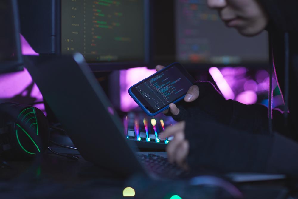 representación de hacker, con una computadora al frente y viendo datos en un celular a su costado derecho